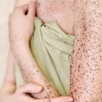 woman applying body exfoliating scrub. natural organic coffee polish on her hands and breast. crop view. home spa concept. skin care and treatment.
